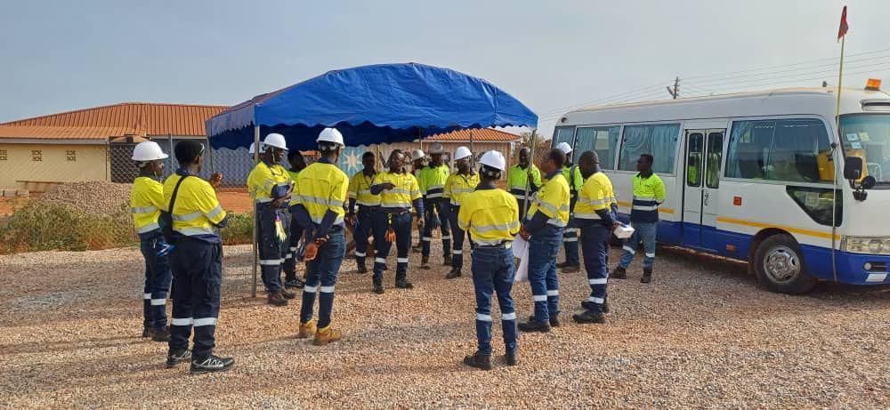 Group of workers in safety gear having a meeting outdoors near a blue tent and a parked bus.