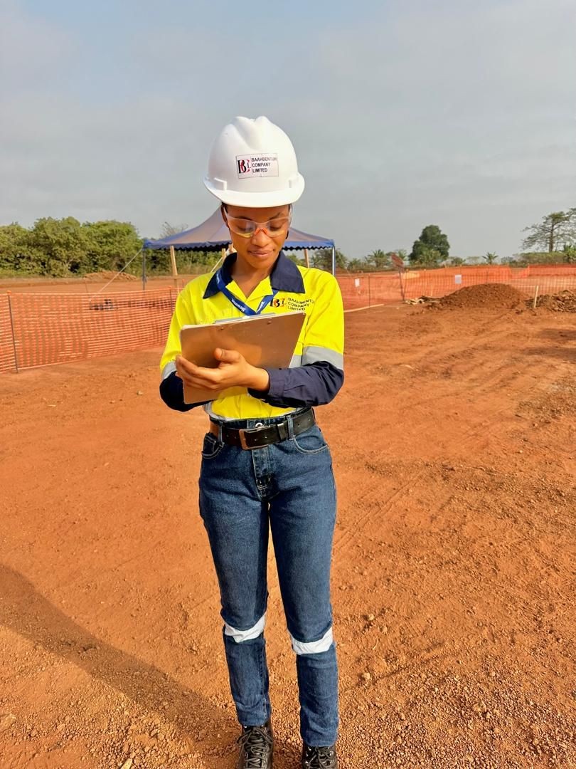 Construction worker in safety gear holding a clipboard at a site with red soil and fenced area.